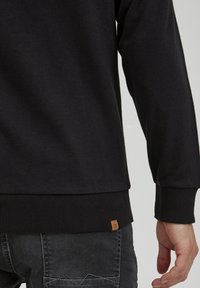 Felpa nera in cotone con polsini e orlo a coste. Presenta un piccolo etichetta logo marrone vicino al fondo, che mostra una forma e una texture aderente.
