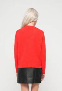 Mulher com cabelo loiro comprido vestindo um suéter vermelho brilhante e uma saia mini de couro preta, voltada para longe, contra um fundo simples.