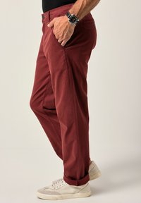 Červené bavlněné chinos kalhoty s volným střihem, s bočními kapsami a ohrnutými nohavicemi, spárované s bílými teniskami. Textura vypadá hladce.