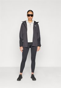 Veste noire résistante aux intempéries avec capuche, associée à un haut gris clair zippé, des leggings noirs et des chaussures de sport noires. Le modèle porte des lunettes de soleil.