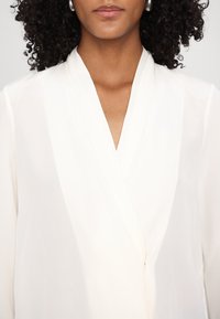Femme en blouse blanche à col en V avec un col subtil et des cheveux bouclés foncés, portant des boucles d'oreilles argentées en anneaux.
