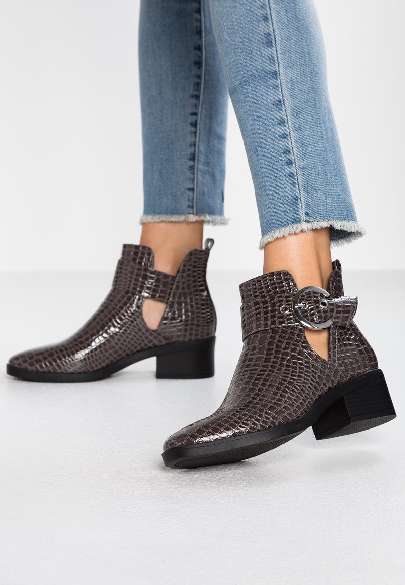 Bottes en cuir marron foncé à motif croco montant à la cheville, avec découpes latérales et grande boucle argentée, portées avec un jean court bleu clair.