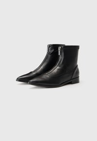 Un par de botas de cuero negras hasta el tobillo con cremalleras plateadas en la parte delantera y punteras puntiagudas sobre un fondo blanco.