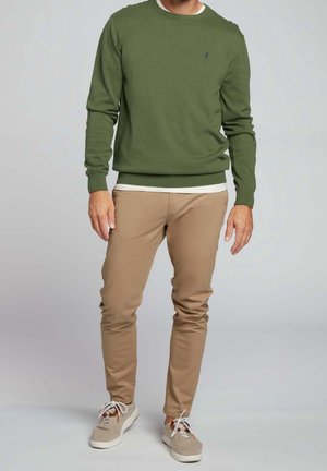 Mężczyzna ubrany w oliwkowozielony sweter z okrągłym dekoltem, beżowe spodnie chino i jasnobeżowe trampki, stojący na tle neutralnym.
