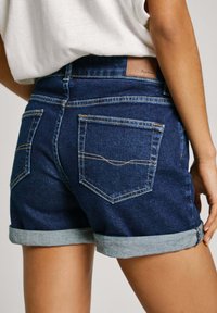Pantalones cortos de mezclilla oscura con puños enrollados, que presentan dos bolsillos traseros y costuras amarillas en contraste. Textura suave con un ajuste clásico.