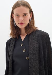 Svart kimono med glittrande textur, lager över en mörk topp med tre runda sköldpaddsknappar. Långa, glänsande silverörhängen.
