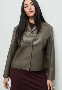 Femme aux longs cheveux noirs portant une veste en cuir olive à boutons par-dessus une robe violet foncé, se tenant devant un fond clair.