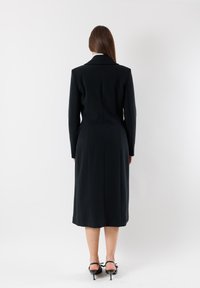 Lungo cappotto nero realizzato in tessuto strutturato, con colletto classico, silhouette aderente e lunghezza al polpaccio, dotato di tasche laterali.