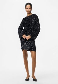 Mulher vestindo um vestido mini de mangas longas preto com lantejoulas, segurando uma pequena bolsa de couro preta e usando sapatos de salto fins pretos.