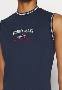 Mörkblå ärmlös topp med hög krage, med vit och röd broderad logotext "TOMMY JEANS NEW YORK CITY" på framsidan.