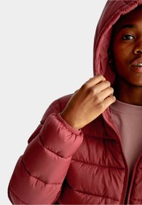 Person iført en rød quilted jakke med hætte, der holder i hættens kant, klædt i en matchende rød skjorte mod en ensfarvet baggrund.