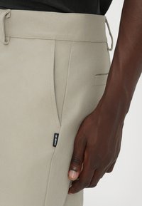 Main à la peau foncée reposant près de la poche avant d’un pantalon beige avec une petite étiquette en tissu noire et blanche sur la couture.