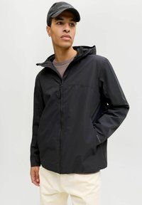 Veste noire à capuche avec fermeture éclair à l'avant, dotée de poignets élastiques et d'une finition mate lisse. Portée sur un haut marron clair et un pantalon crème.