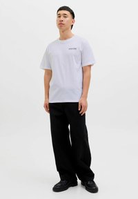 Vaaleanharmaa puuvillainen t-paita lyhyillä hihoilla, jossa on pieni musta logo rinnassa; yhdistettynä väljiin mustiin housuihin ja mustiin kenkiin.