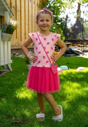 Ein lächelndes Mädchen in einem rosafarbenen Top und Rock mit Erdbeermuster steht auf dem Gras mit den Händen in den Hüften, trägt eine Erdbeertasche und ein Haarband.