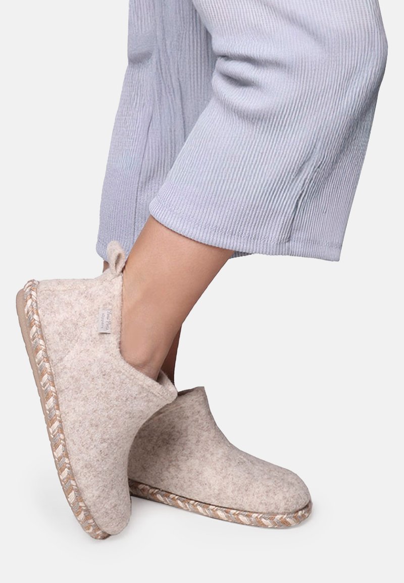 Pieds chaussés de chaussons beige clair en laine avec bordure tissée sur la semelle, assortis à un pantalon court côtelé gris clair.