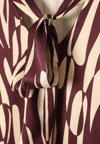 Blouse bordeaux et crème avec un détail noué au niveau du col, présentant une texture lisse et des motifs de feuilles abstraites et allongées.