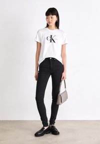 Sieviete stāv pie baltām sienām, ģērbusies baltā Calvin Klein logo t-kreklā, melnās šaurās biksēs, melnās mokasīnās un turētā pelēkā rokassomā.