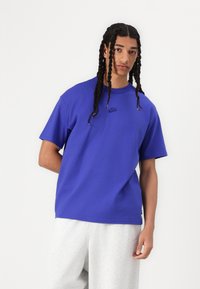 T-shirt en coton bleu à manches courtes, col rond et petit logo Nike noir sur la poitrine. Porté avec un pantalon de survêtement gris.