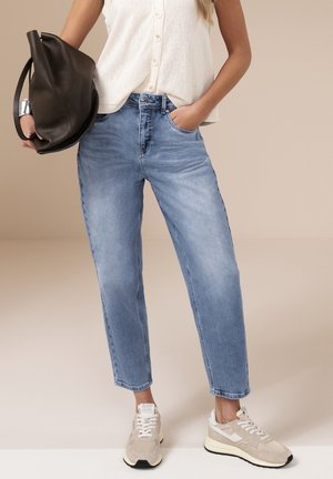 Kvinde iført lyseblå højtaljede jeans, hvid ærmeløs bluse med knapper, beige sneakers, sølv manchetarmbånd, holder stor brun lædertaske.