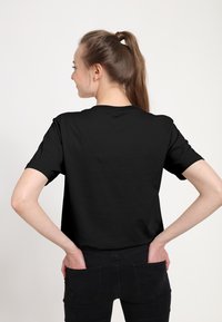 Žena s hnědým copem, která má na sobě jednoduché černé tričko a černé džíny, se rukama v zadních kapsách, otočená zády k černobílému pozadí.