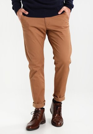 Hombre con pantalones marrón camello remangados, botas de cuero marrón oscuro con cordones y suéter azul marino, con las manos en los bolsillos, de pie sobre un fondo blanco.
