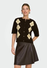 Cardigan en maille marron avec motif de losanges crème, manches courtes, encolure ronde et boutons sur le devant, associé à une jupe en cuir marron ajustée.