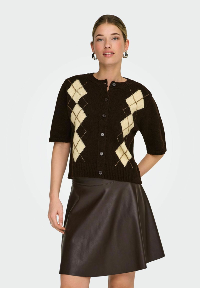 Cardigan en maille marron avec motif de losanges crème, manches courtes, encolure ronde et boutons sur le devant, associé à une jupe en cuir marron ajustée.