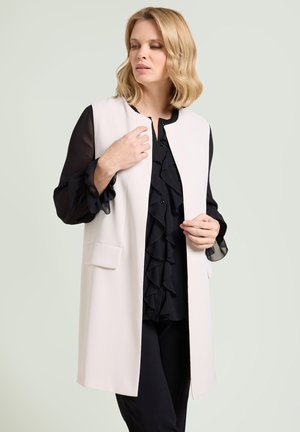 Vrouw met schouderlang blond haar, die een zwarte blouse met ruches en een mouwloos wit vest draagt, kijkt opzij.