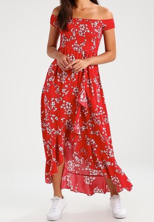 Femme portant une robe longue fleurie rouge à épaules dénudées, associée à des baskets blanches, se tenant devant un fond uni.