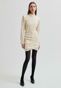 Kobieta w kremowej mini sukience ozdobionej cekinami z długimi rękawami, czarnych rajstopach oraz czarnych szpilkach z wycięciami.