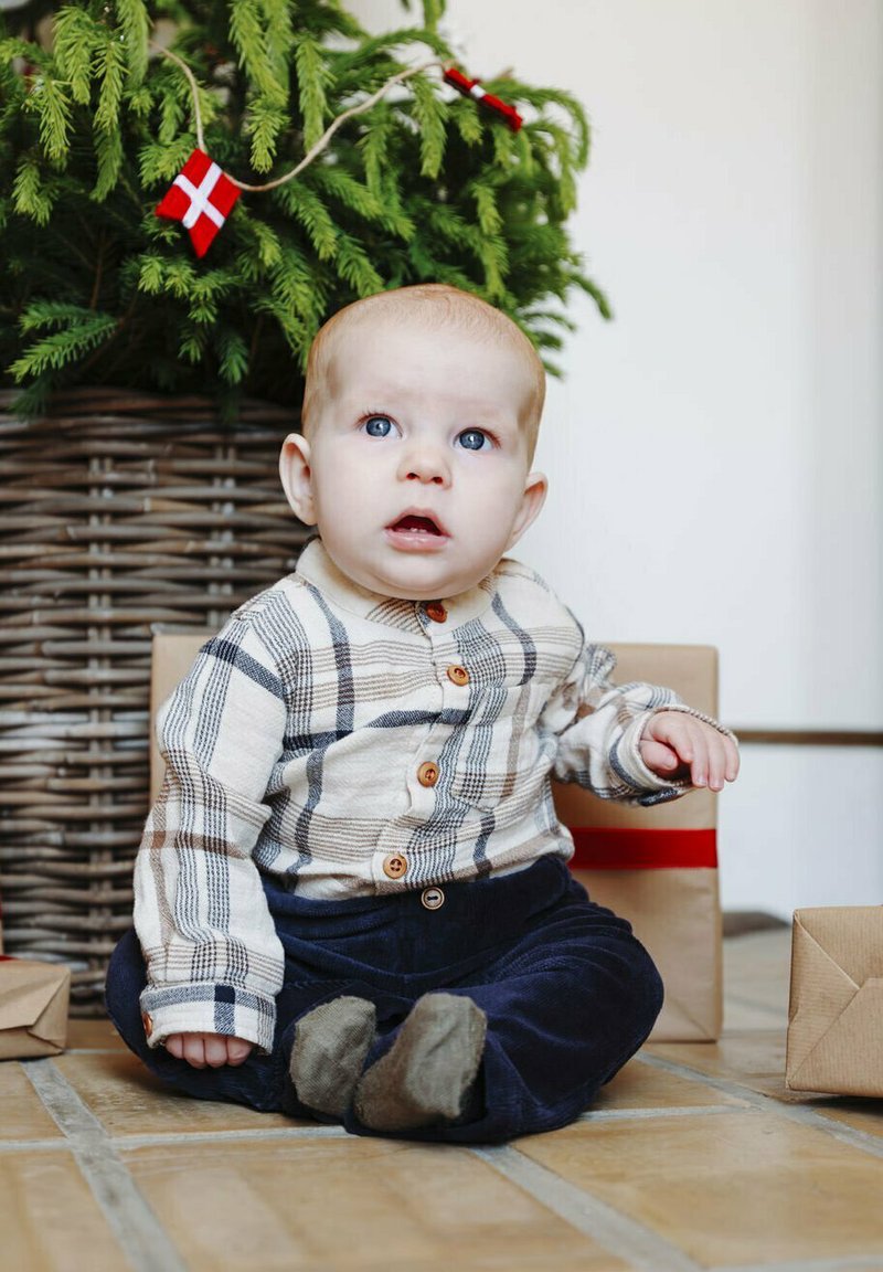 Camicia a scacchi beige e marrone con bottoni, pantaloni di velluto a coste scuri, seduto su un pavimento piastrellato con regali incartati e un albero di pino verde sullo sfondo.
