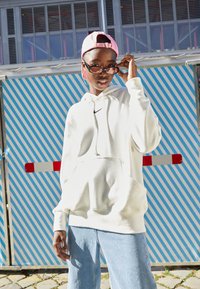 Hvit hettegenser med frontlomme og snøring, kombinert med lyseblåjeans. Modellen har på seg en rosa baseballcaps og briller. Stripet bakgrunn.