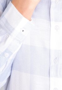 Lyseblå og hvid ternet skjorte lavet af tekstureret stof. Har rund krave og små hvide knapper på fronten og ærmerne.