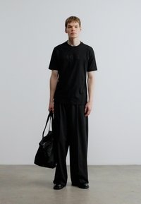 Fekete pamut póló domború logóval, széles fekete nadrággal párosítva. A modell fekete táskát visel és fekete cipőt hord.