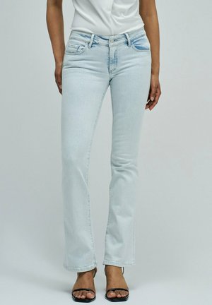 Personne portant un jean bleu clair à jambes droites et des sandales noires à talons ouvertes, debout devant un fond clair uni.