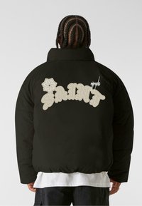 Giubbotto imbottito nero con vestibilità oversize, caratterizzato da testo "SAINT" color crema e design floreale sul retro, e colletto alto.