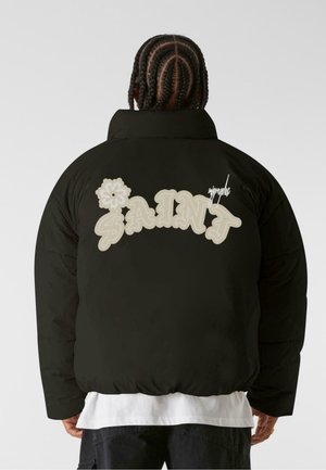 Czarna kurtka puchowa o oversize'owym kroju, z kremowym napisem "SAINT" i kwiatowym wzorem z tyłu oraz wysokim kołnierzem.