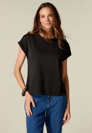 Kvinde med langt brunt hår iført en sort kortærmet top, blå jeans og et sort armbånd på venstre håndled, stående mod en beige baggrund.