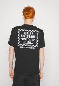 Černé bavlněné tričko s velkým bílým potiskem na zádech, který zobrazuje "REPLAY SPEEDSHOP" a kostkované akcenty.
