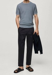 Svetle modrý texturovaný svetr s krátkým rukávem, tmavé zužované kalhoty, černé sandály. Drží tmavý svetr. Jednoduchý, funkční design.