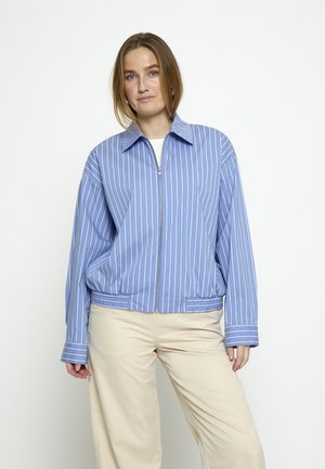 Žena sa kosom do ramena, odjevena u plavu i bijelu prugastu jaknu na patentni zatvarač i bež hlače, stoji nasuprot bijeloj pozadini.