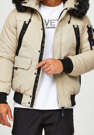 Mann trägt beige gesteppte Jacke mit schwarzen Reißverschluss-Details und fellbesetzter Kapuze über weißem Hemd und schwarzer Hose, zeigt auf die Vorderseite der Jacke.