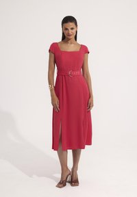 Czerwona sukienka z kwadratowym dekoltem, rękawami typu cap i paskiem w talii z okrągłą klamrą. Szczeliny na końcu podkreślają design.