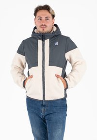 Giacca con cappuccio grigia e crema con zip frontale, caratterizzata da una texture in fleece e due tasche anteriori. Logo del marchio sul lato sinistro in alto.