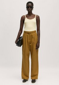 Crème mouwloze top gecombineerd met hooggerdealde, mosterdkleurige wijde broek. Model houdt een zwarte clutch vast. Accessoires omvatten zwarte zonnebril en creolen.