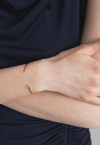 Brățară din metal cu finisaj auriu, purtată pe încheietura unei mâini, având un design elegant, minimalist, cu un finisaj neted și detalii accent subtile.