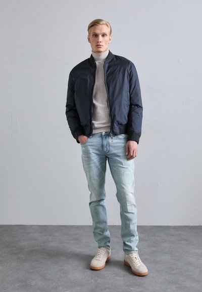 Marinblå bomberjacka med en subtil textur, ljusgrå polo, slitna ljusblåjeans och beige sneakers med gummisulor.