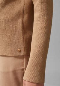 Beżowy sweter o teksturze z dopasowanym krojem. Posiada okrągły dekolt i złoty guzik akcentowy przy dolnym brzegu. Gładkie wykończenie materiału.