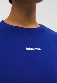 Tricou albastru Lululemon cu guler rotund. Prezintă un design simplu și un logo alb centrat în zona pieptului. Textură moale.